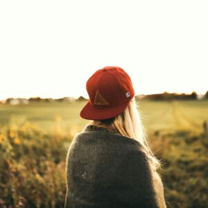 Back view of a woman in a red cap and sweater standing in a field during sunset.